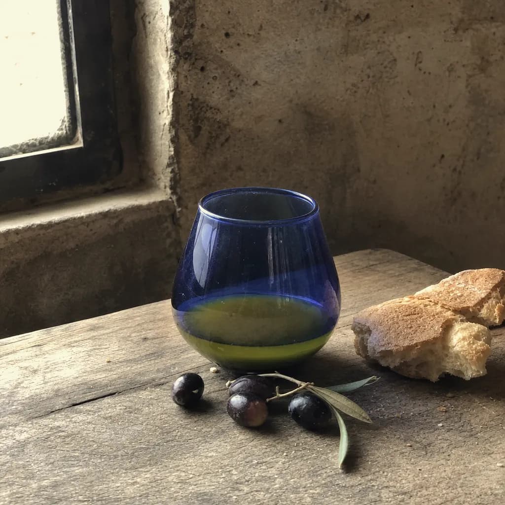 Freshly made olive oil in a blue tasting glass beside olives and bread