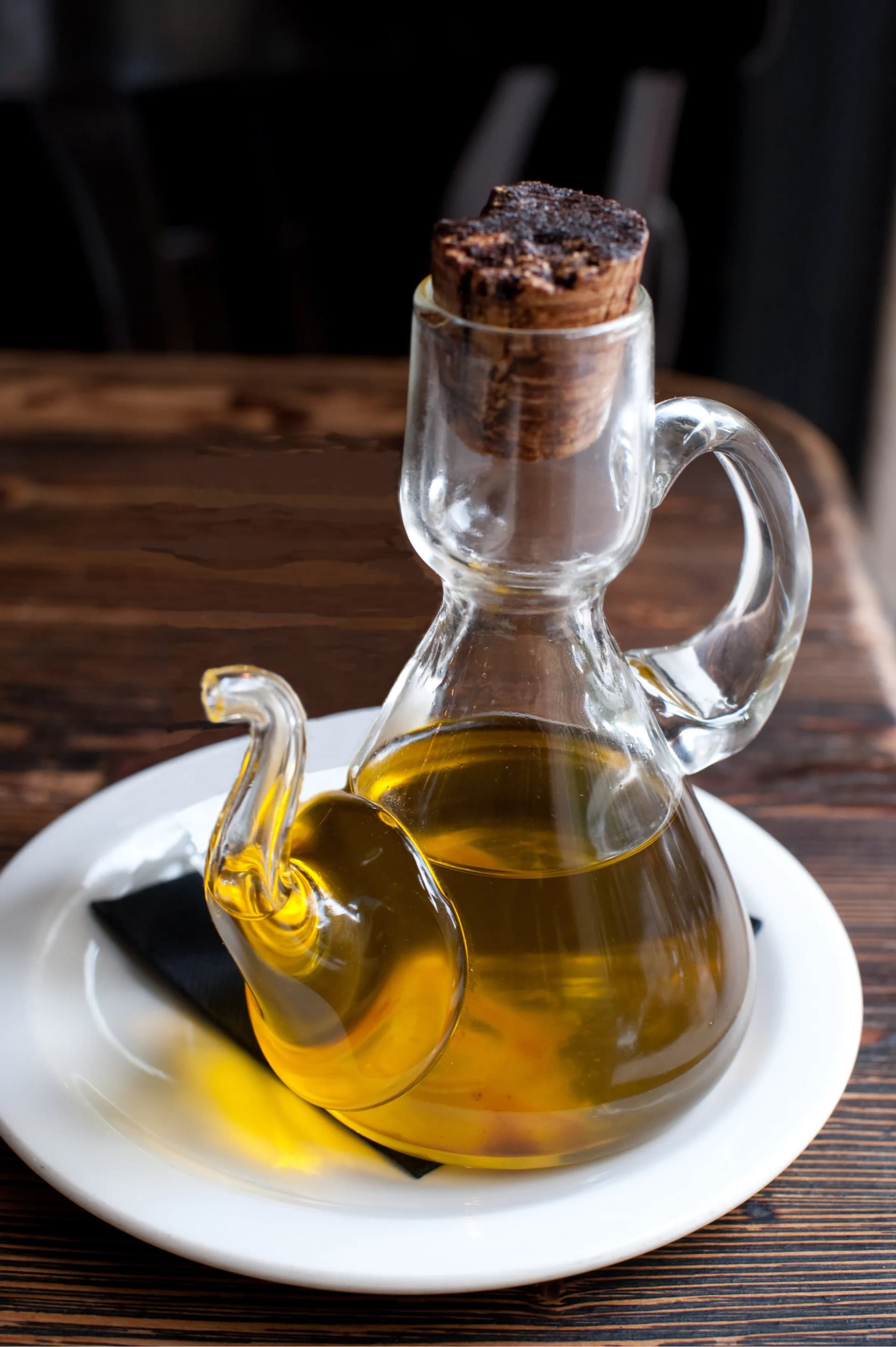 A glass oil cruet served on a plate on a dark wooden table