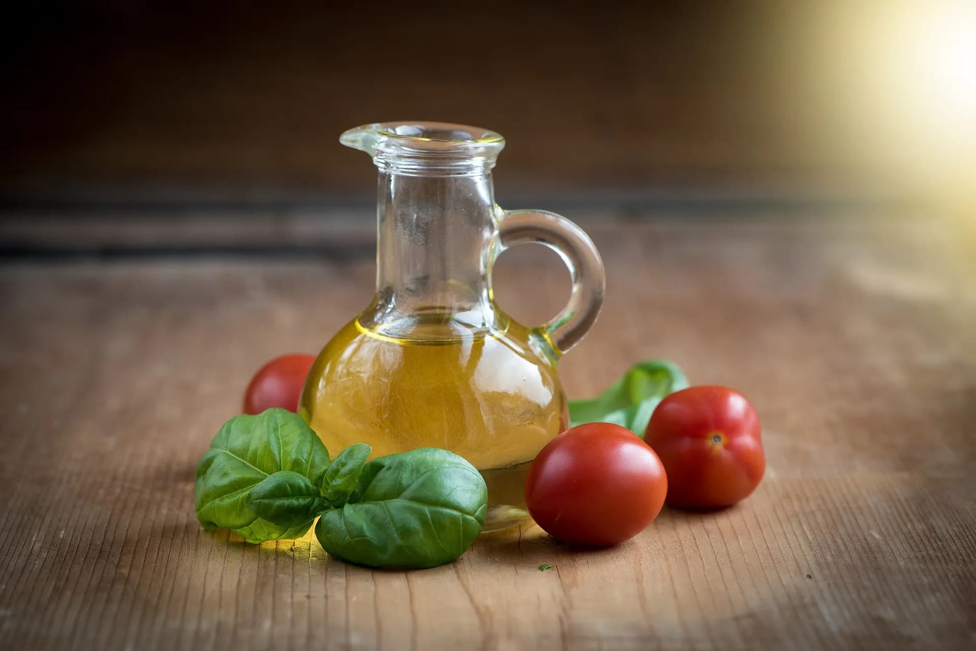 A glass oil cruet on wood, with basil and tomatoes beside it