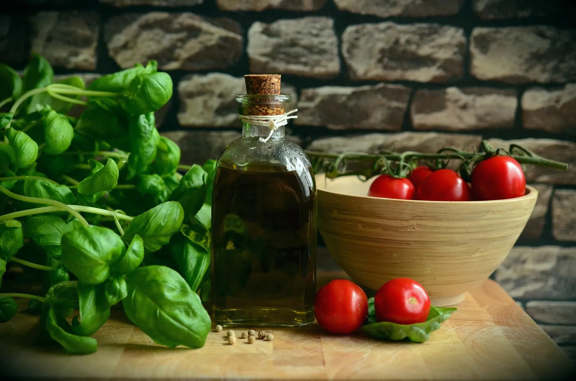 A bottle of oil beside basil and tomatoes, with a stone wall behind